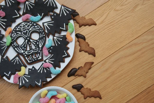 Halloween cookies in shape of skull and bat with colorful worm candies on a plate on wooden background. Halloween sweet food