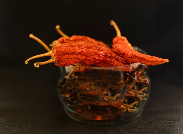 A bunch of dry long red chillies placed in a glass bowl with a black backdrop.
