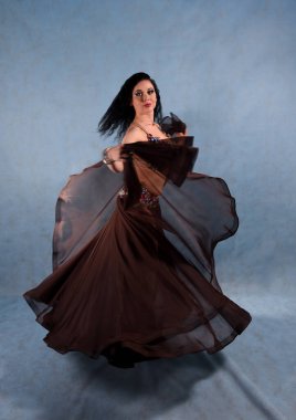 Oryantal elbiseli güzel bir dansçı stüdyoda mavi arka planda dans ediyor kahverengi etek ve elbisesini sallıyor.