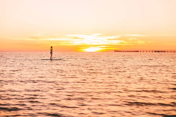 Surf woman rowing on the surfboard in ocean at sunset.