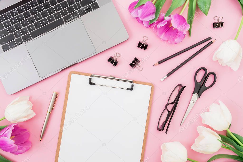 Laptop on pink background with clipboard, tulips flowers, glasses and accessories. Flat lay. Top view. Freelancer office concept.