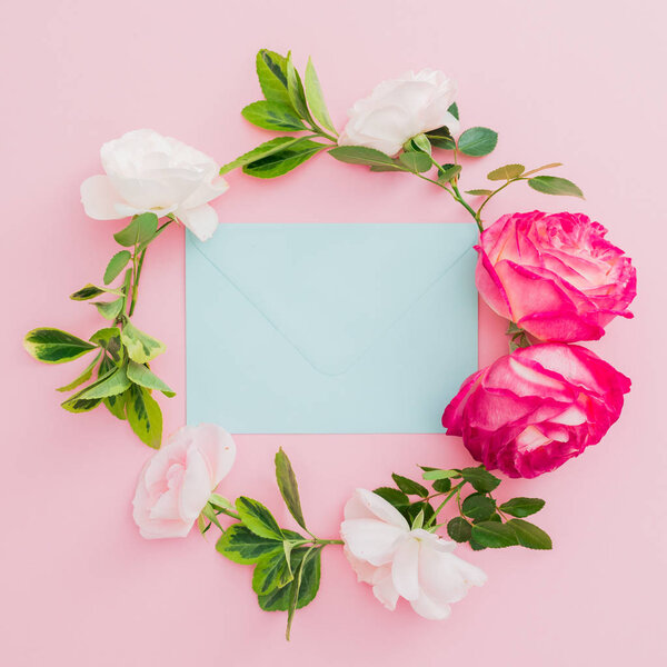 Wreath floral frame with white and red roses flowers and blue envelope on pink background. Flat lay, top view.
