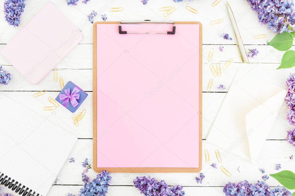 Feminine work place composition with clipboard, notebook, lilac flowers, gift box and accessories on rustic background. Flat lay, top view.