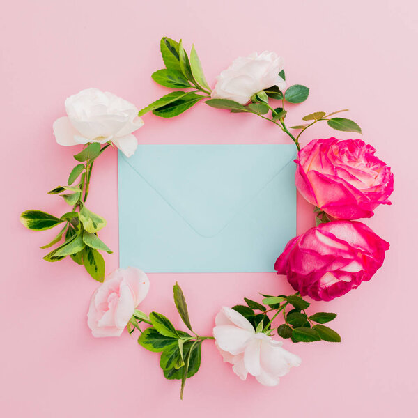 Floral frame with roses flowers and blue envelope on pink background. Flat lay, top view.