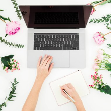 Dizüstü bilgisayarda çalışan kız ve günlüğüne yazmak. Office workspace eller, dizüstü bilgisayar, pembe çiçekler ve beyaz masada günlüğü ile. Üstten görünüm. Düz yatıyordu.
