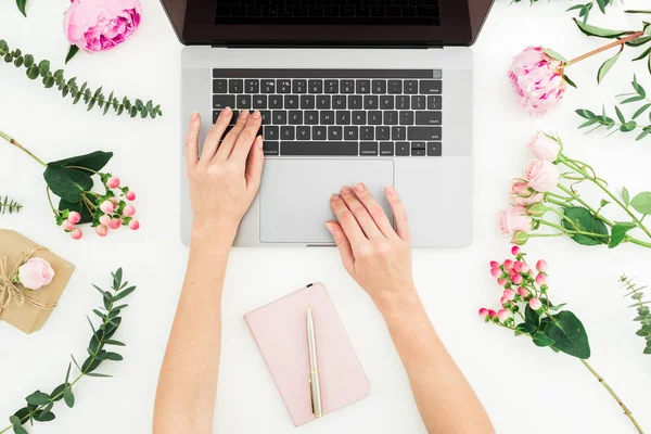 Girl typing on laptop. Office workspace with female hands, laptop, notebook and pink flowers on white background. Top view. Flat lay.