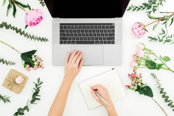 Woman work with laptop. Workspace with female hands, laptop, notebook and pink flowers on white background. Top view. Flat lay.