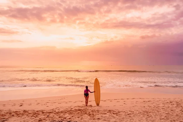 日没や日の出のビーチでサーフボードを持つサーファーの女性 — ストック写真
