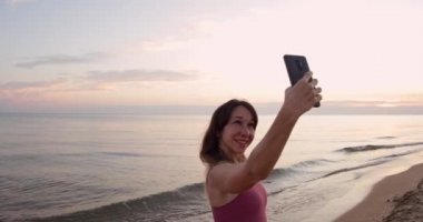Çekici bir kadın elinde telefonla sahilde gün batımını ya da güneşin doğuşunu çekiyor.