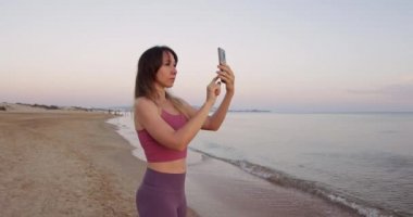 Çekici bir kadın elinde telefonla sahilde gün batımını ya da güneşin doğuşunu çekiyor.