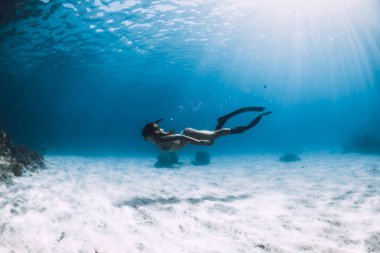 Yüzgeçli sporcu bir kadın okyanusun altında mercanların yanında kumlu zeminde süzülüyor.