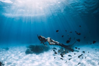 Yüzgeçleri olan serbest dalgıç genç bir kadın şeffaf okyanusta tropik balıklarla kumlu zeminde süzülüyor.