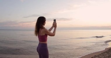 Akıllı telefonlu çekici bir kadın, gün batımını ya da deniz sahilinde gün doğumunu çekiyor.