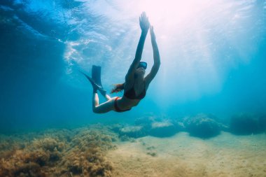 Bikinili zayıf bir kadın güneş ışınlarıyla mavi denizde süzülüyor. Suyun altında yüzgeçleriyle serbest dalış