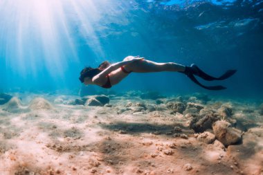 Bikinili serbest yüzen zayıf kadın mavi denizde ve güneş ışınlarında süzülüyor. Okyanusun altında yüzgeçleriyle serbest dalış