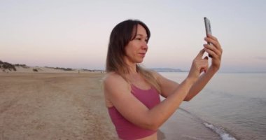 Akıllı telefonuyla ayakta duran ve gün batımını ya da kumsalda gün doğumunu çeken çekici bir kadın.