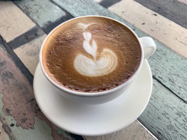 Beyaz bardakta kahve ve çay tabağı. Pastel ahşap bir masa üzerinde güzel bir Latte Art ve üst manzarada klasik bir desen..