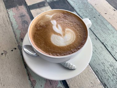 Bardaktaki kahvenin beyaz bir fincan tabağı ve pastel ahşap bir masa üzerinde güzel bir Latte Art ile bir kaşığı ve üst manzarada klasik desenleri var..
