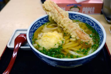 Tempura udon, yumurta ve chikawa ile berrak bir çorba, üzerinde kıymalı soğan, siyah bir tepsiye yerleştirilmiş kase yanında bir kaşık ile.