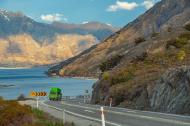 Yeşil araba tepeden kıvrımlı bir yola doğru koşuyor. Akşamları denizi ve dağları ve mavi gökyüzünü görmek Hawea Gölü, Wanaka Otago, Yeni Zelanda.