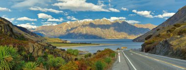 Hawea Gölü 'ndeki güzel geniş ekran panoramik manzara, denize giden yokuş aşağı yollar ve akşamları dağlar ve Wanaka Yeni Zelanda' da bulutlu mavi gökyüzü..