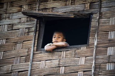 Chiang Mai / Tayland - 16 Ocak 2016: Sert tenli çocuk Gövdesi fakir görünüyordu, çocuk ahşap evin penceresine tünemişti. Duvarlar örülmüş bambudan yapılmış, sıkılmış, mutsuz ve umutsuz görünüyorlar.