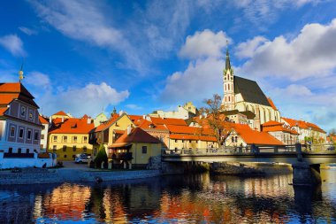 Cesky Krumlov 'daki Vltava Nehri manzarası ve St. Vitus Kilisesi, öğleden sonra mavi gökyüzü ve güzel bulutlarla. Köprüde turistler var..
