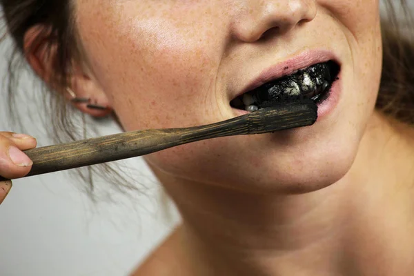 Young woman brushing her teeth with a black tooth paste with active charcoal, and black tooth brush on white background for Teeth whitening — Stock Photo, Image