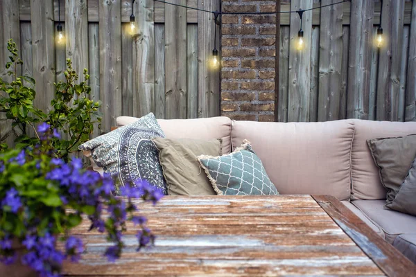 Стильная садовая мебель, диван с подушками и лампами, подвешивание лампочек, уютный современный уголок на террасе — стоковое фото