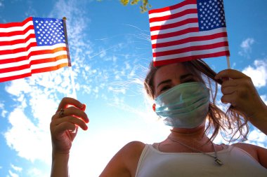 Amerikan bayrağını mavi gökyüzünde tutan genç bir kadın güneş ışığı ve güvenlik maskesiyle Covid-19 Amerika Birleşik Devletleri için