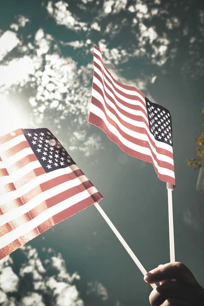 Χέρι κρατώντας δύο αμερικανικές σημαίες στον γαλάζιο ουρανό με φόντο το φως του ήλιου, κυματίζει σημαία για τις Ηνωμένες Πολιτείες της Αμερικής close-up — Φωτογραφία Αρχείου