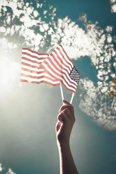 Hand holding two american flags on the blue sky with sunlight background, waving flag for United States of America close-up