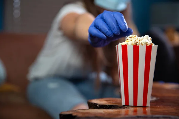 Mujer con máscara de seguridad y guantes para Covid-19, coronavirus y cuarentena viendo televisión en casa con cubo de armario a rayas rojas y blancas con palomitas de maíz. Cine y concepto de salud — Foto de Stock