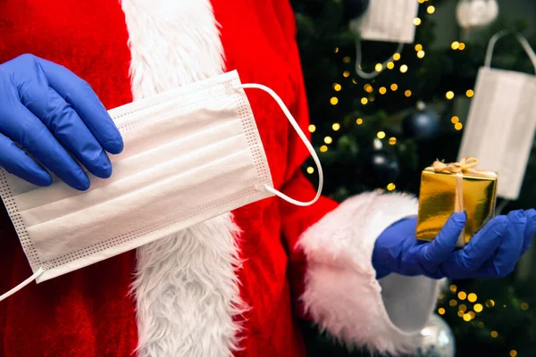 Weihnachtsmann mit Handschuhen und Mundschutz für Coronavirus am Weihnachtsbaum mit goldenem Geschenk, Covid-19 und Weihnachtssicherheitskonzept — Stockfoto