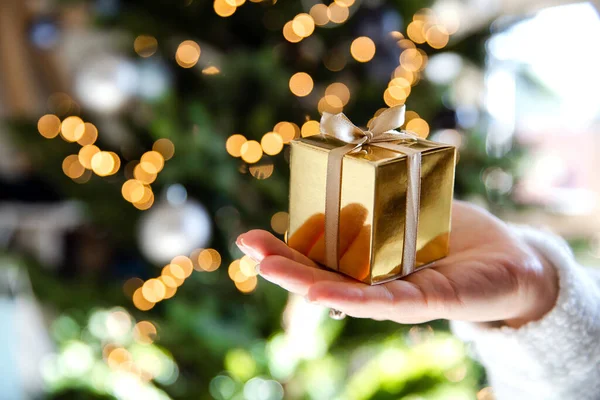 Покриття руки, що тримає золоту коробку з різдвяним деревом і вогні на задньому плані, сьогоденні і різдвяні свята концепції — стокове фото