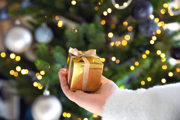 Покриття руки, що тримає золоту коробку з різдвяним деревом і вогні на задньому плані, сьогоденні і різдвяні свята концепції — стокове фото