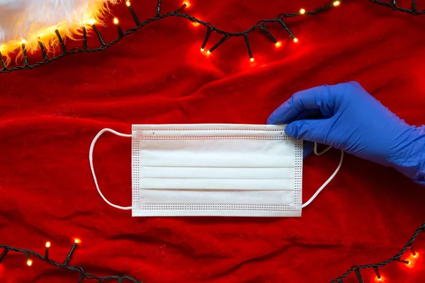 Dua tangan dengan sarung tangan pelindung memegang topeng wajah medis dengan latar belakang merah dengan lampu Natal, konsep untuk Covid-19 dan Natal, latar belakang coronavirus — Stok Foto
