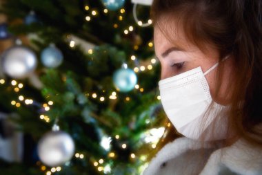 Koruyucu yüz maskesi takan ve arka planda Noel ağacı, koronavirüs ve Noel konsepti olan Covid-19 için üzgün görünen genç bir kadının portresi.