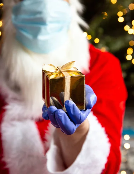 Der Weihnachtsmann hält eine goldene Geschenkbox mit Schutzhandschuhen und Schutzmaske gegen Coronavirus, Covid-19 und Weihnachtskonzept in der Hand — Stockfoto