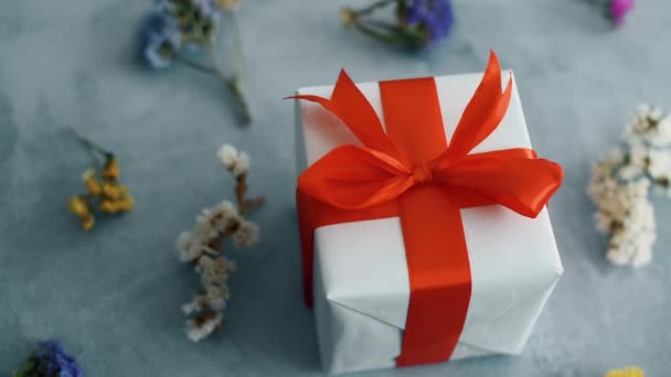 赤いシルクのリボンが乾いた色の花でテクスチャ表面に回転する白いギフトボックス クリスマス ブラックフライデー その他の休日のためのギフトボックス 4K映像をクローズアップ — ストック動画
