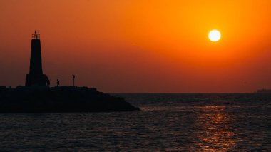 amazing sunset in puerto banus, marbella. orange and soft orange in the sky with a calm sea, and the light house in the scene clipart