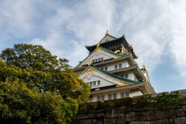 Yeşil geleneksel çatıları ve altın aşermeleri olan Osaka Şatosu görkemli ana kulesi, arka planda mavi gökyüzü ve ağaçlar, sonbahar, Japonya.