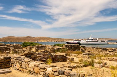 DELOS, Yunanistan. Yunanistan 'ın Delos Adası' ndaki bir körfez manzarasında, Mykonos 'tan demirlemiş turist gemileri ve ön planda antik şehir harabeleri ve sütunları yer alıyor..