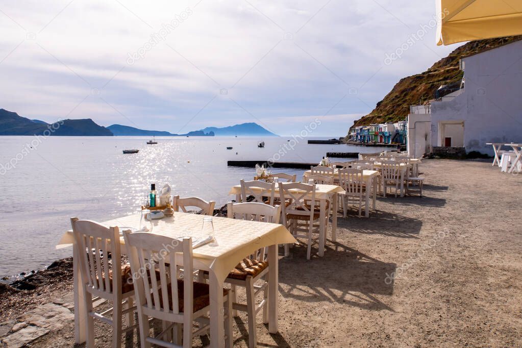 White dining tables on the sea shore with impressing sea view and Klima fishermen village in the background on Milos Island, Greece.