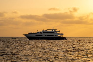 Kızıl Deniz üzerinde turuncu gün batımlı, bulutlu gökyüzü ve sakin deniz olan bir dalış teknesinin karanlık silüeti..