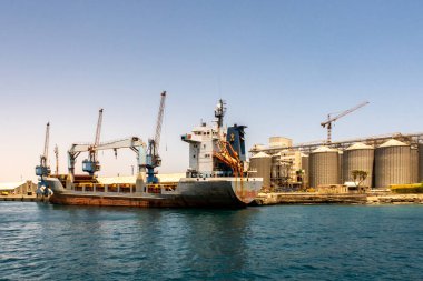 Port Sudan limanında büyük bir konteyner gemisi kargoyla yükleniyor. Arka planda büyük iskele vinçleri ve sıra sıra tahıl siloları.