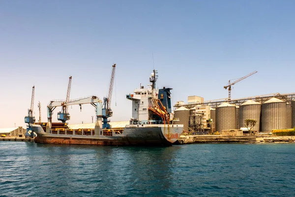 Port Sudan limanında büyük bir konteyner gemisi kargoyla yükleniyor. Arka planda büyük iskele vinçleri ve sıra sıra tahıl siloları.