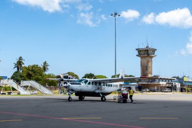 Zanzibar, 28/09/18. Zanzibar International Airport apron with Cessna 208 Caravan of Coastal Air local airline parked and staff loading luggage, with control tower and terminal in the background. clipart