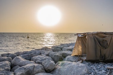 Taştan bir iskelede duran eski halılardan yapılmış geçici kumaş çadırı. Arka planda günbatımı var..