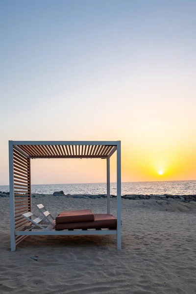 Empty double sun bed on the sandy beach in Dubai, with orange sunset in the background and calm sea, portrait view.